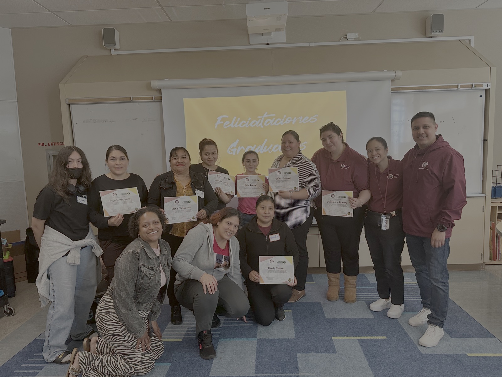 Parent graduates holding certificates in front of a Felicitaciones Graduadas screen